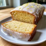 A slice cut from a glazed lemon loaf, showing the moist crumb and topped with lemon zest.