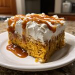 A moist slice of pumpkin poke cake topped with whipped cream and drizzled generously with caramel sauce.