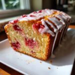 A close-up of a moist strawberry bread loaf, studded with red berries and drizzled with white icing.