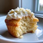 Close-up of a vanilla cupcakes with a bite taken out, showing the moist crumb and white vanilla frosting.