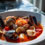 A close-up, appetizing shot of a bowl of rich tomato-based seafood soup featuring mussels, clams, shrimp, and scallops.