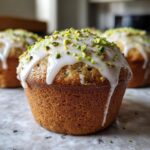 A close-up of a freshly baked pistachio muffin topped with white glaze and chopped green pistachios.