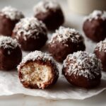 Close-up of chocolate covered coconut balls, one is bitten open revealing the shredded coconut filling.