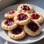 A close-up of several buttery raspberry thumbprint cookies piled on a white plate, featuring bright red jam centers.