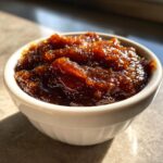 A close-up of thick, dark, glistening homemade apple butter served in a small white ramekin.