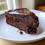 A decadent slice of flourless chocolate cake with a shiny dark chocolate ganache topping on a white plate.