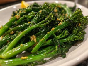 Close-up of bright green roasted broccolini stalks seasoned with visible bits of garlic and oil, served on a white plate.