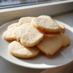 A pile of freshly baked, light golden sugar cookies, some heart-shaped, resting on a white plate near a window.