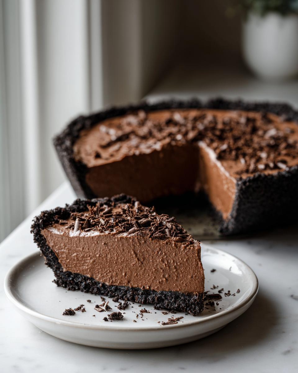 A decadent slice of chocolate mousse pie with a dark crust sits on a white plate, with the rest of the chocolate mousse pie in the background.