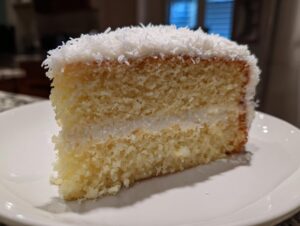 Close-up of a moist slice from a 3-layer coconut cake recipe, showing fluffy layers and shredded coconut topping.