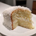 Close-up of a moist slice of three-layer coconut cake recipe covered in shredded coconut.