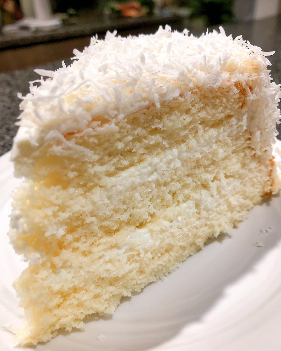 Close-up of a moist slice from a 3-layer coconut cake recipe, covered in white frosting and shredded coconut.