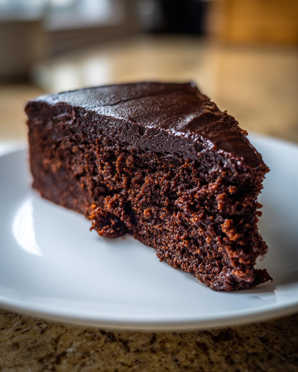 A close-up of a moist slice of flourless chocolate cake topped with shiny chocolate ganache on a white plate.