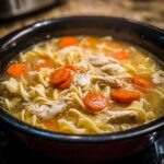 Close-up of steaming slow cooker chicken noodle soup with egg noodles, shredded chicken, and bright orange carrots.