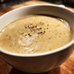 Close-up of a white bowl filled with thick, smooth cream of chicken soup, garnished with pepper and herbs.