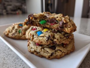 A stack of three chewy kitchen sink cookies, one broken open to show chocolate chunks and colorful M&Ms inside.