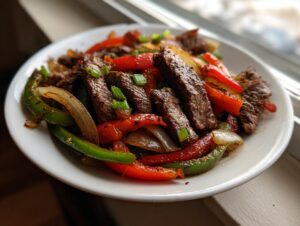 Close-up of juicy sliced steak fajitas mixed with grilled red and green bell peppers and onions on a white plate.