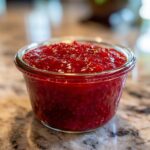 Close-up of bright red strawberry freezer jam filling a small glass jar on a granite countertop.