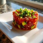 A beautifully molded tower of vibrant red tuna tartare mixed with avocado chunks, topped with sesame seeds and green onions.