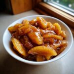 A white bowl filled with glistening, spiced apple pie filling, showcasing thick apple slices coated in cinnamon sauce.