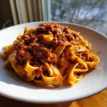 A close-up of fresh tagliatelle pasta topped generously with rich, hearty bolognese sauce, served in a white bowl.