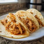 Three soft corn tortillas filled with shredded, saucy chicken and topped with crumbled white cheese, ready to eat as chicken tacos.