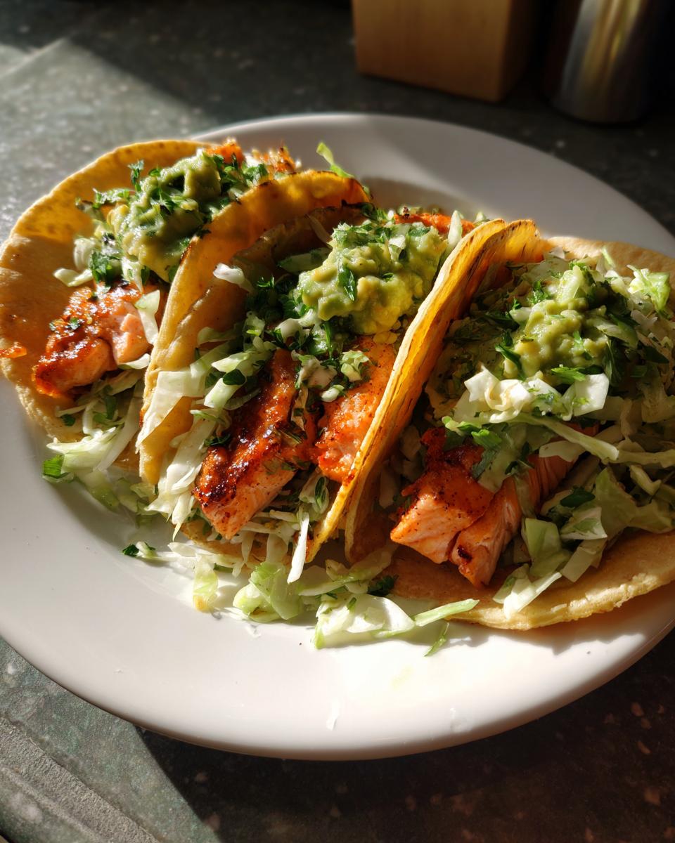 Three fully loaded salmon tacos featuring grilled salmon, shredded lettuce, and a generous dollop of guacamole on corn tortillas.