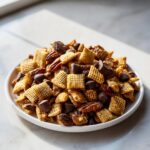 A close-up of a white plate piled high with toffee chex mix recipe ingredients, including Chex cereal, pecans, and chocolate chunks.