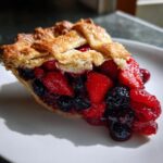 Close-up of a juicy slice of triple berry pie showing strawberries, blueberries, and raspberries under a golden lattice crust.