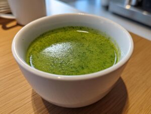 Close-up of a small white bowl filled with vibrant green, frothy spinach soup resting on a wooden surface.