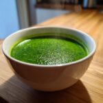 A close-up of a white bowl filled with vibrant, steaming hot spinach soup resting on a wooden surface.