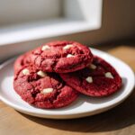 A close-up of three rich, deep red velvet cookies studded with white chocolate chips on a white plate.