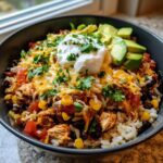 A close-up of a vibrant chicken burrito bowl topped with shredded cheese, sour cream, avocado slices, and cilantro.