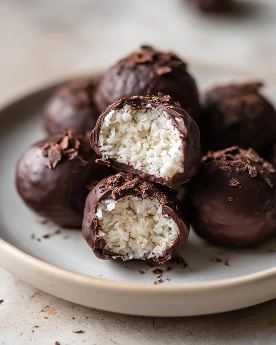 A close-up of homemade almond joy truffles, one cut open revealing the shredded coconut filling inside the dark chocolate shell.