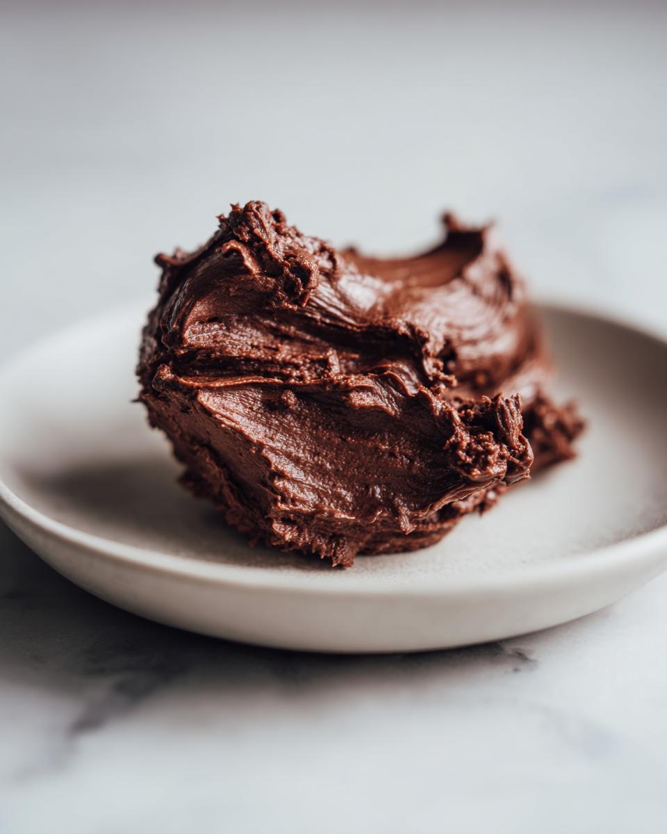 A close-up of a dollop of rich, dark chocolate frosting on a small white plate.