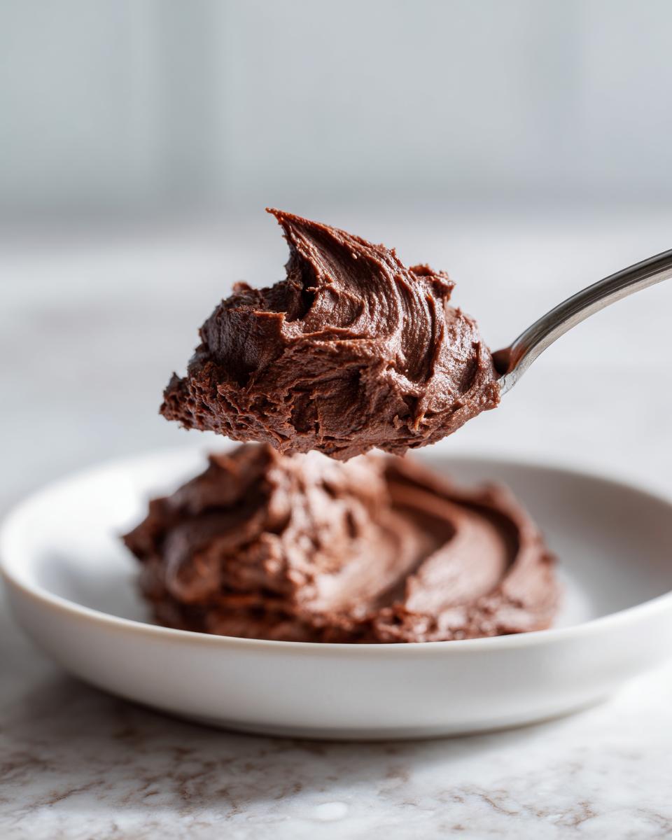 A spoonful of rich, dark chocolate frosting being lifted above a white bowl filled with more chocolate frosting.
