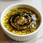 Close-up of a white ramekin filled with vibrant olive oil bread dip, topped with herbs, garlic, and a balsamic vinegar swirl.