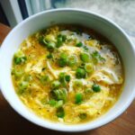 A close-up view of a bowl of savory egg drop soup, featuring wispy cooked egg ribbons and topped generously with bright green sliced scallions.
