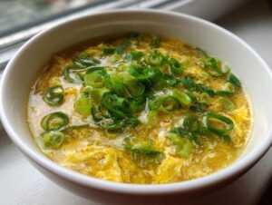 Close-up of a white bowl filled with homemade egg drop soup, featuring wispy egg ribbons and fresh green scallions.