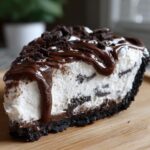 A decadent slice of ice cream pie featuring a dark cookie crust, white creamy filling with cookie pieces, topped with chocolate fudge and crumbles.