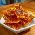 Close-up of shiny, amber-colored pieces of homemade peanut brittle recipe stacked in a small white dish.