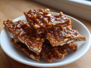 Close-up of broken pieces of glossy, amber peanut brittle recipe stacked in a small white bowl.