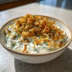 A creamy, green-flecked pickle dip topped generously with crispy fried onions and fresh dill.
