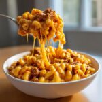 A spoonful of cheesy, saucy taco pasta being lifted from a white bowl, showing extreme cheese pull.