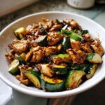 A close-up, appetizing shot of chicken zucchini stir fry coated in a dark, savory sauce served in a white bowl.