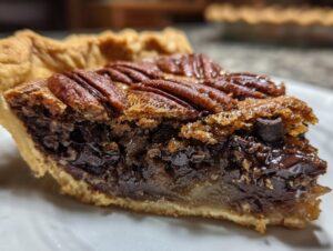 Close-up of a rich slice of chocolate bourbon pecan pie showing the flaky crust, gooey filling, and whole pecans on top.
