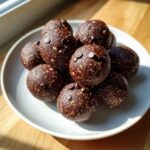 A close-up of several rich, dark chocolate chip protein balls stacked on a white plate in bright sunlight.