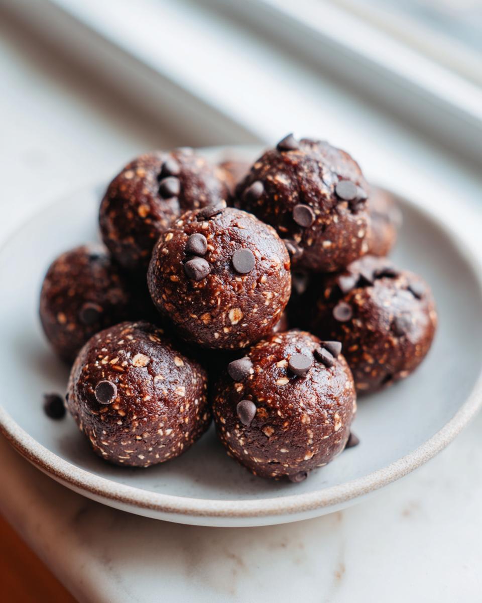 A close-up of several rich, dark chocolate chip protein balls piled high on a light gray plate.
