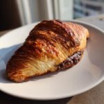 A golden brown, flaky chocolate croissant resting on a light-colored plate, showing melted chocolate oozing out.