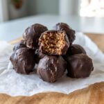 A stack of chocolate-covered pecan pie balls, with one showing a bite taken out to reveal the textured filling.